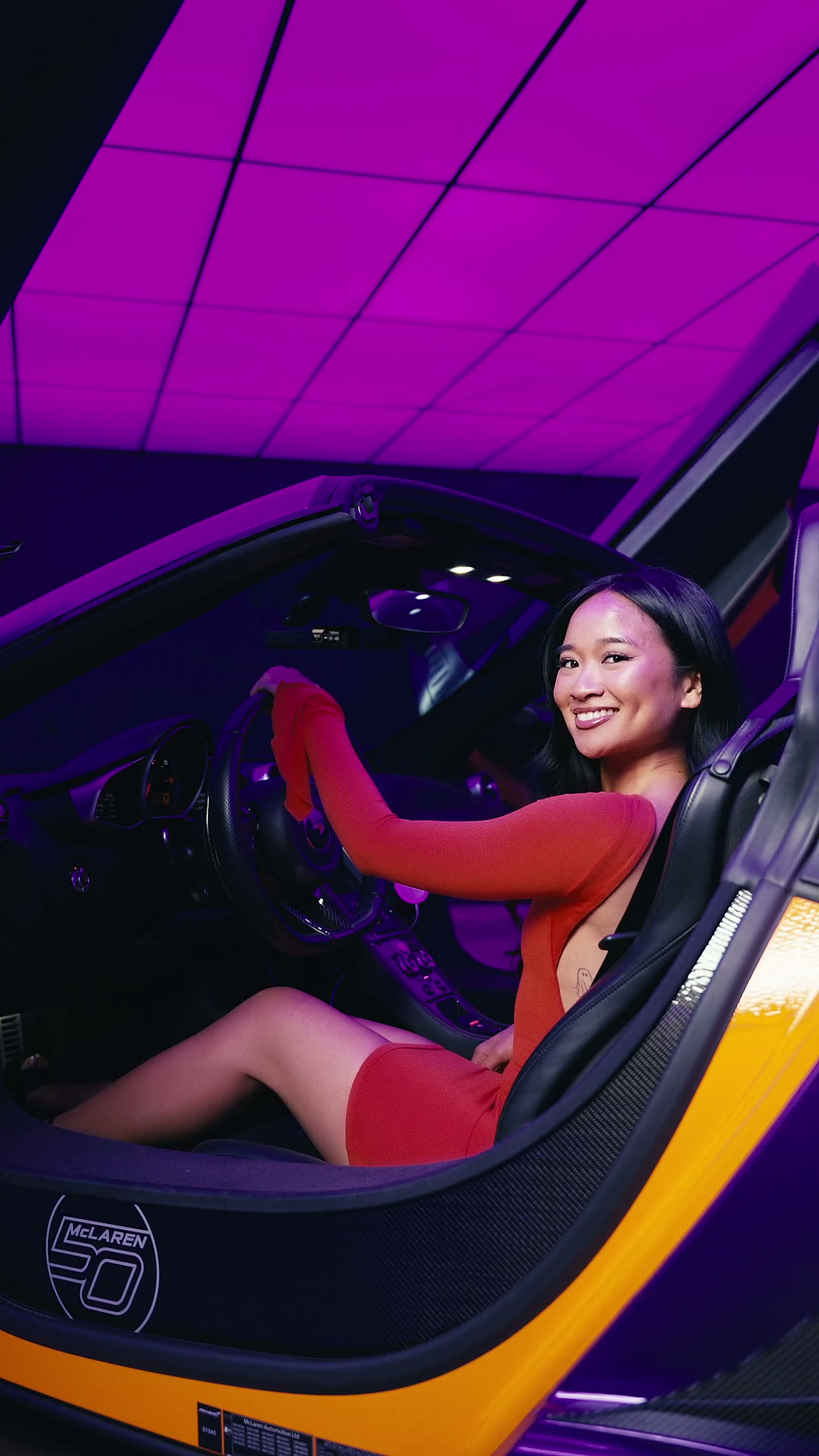 Kamryn Hernandez in a red dress seated at the wheel of a McLaren under magenta studio light